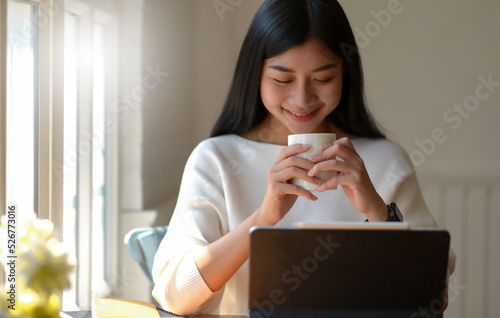 Asian woman holding a cup of coffee and looking down at laptop.