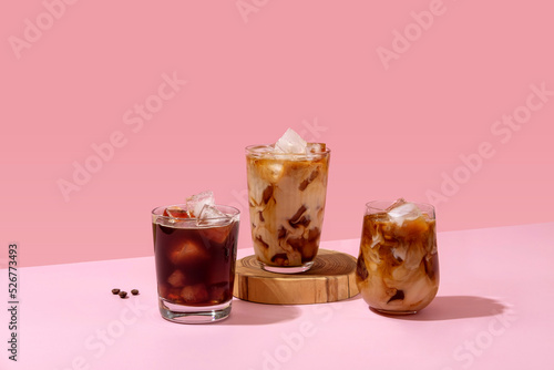 Ice coffee in a tall glass with cream poured over and coffee beans. Set with different types of coffee drinks on a pink table.