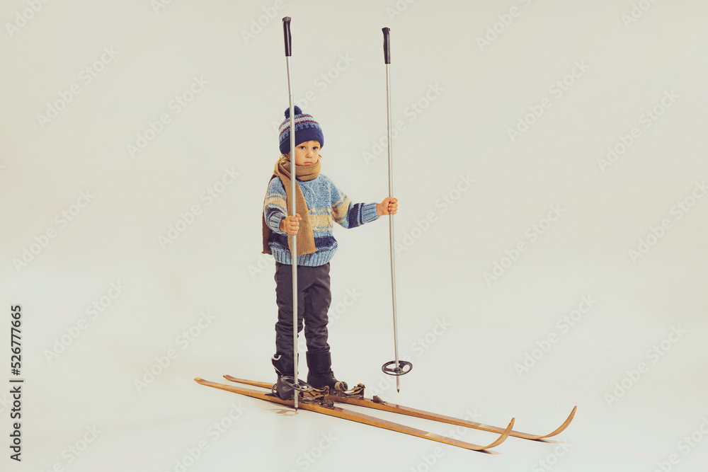 Portrait of little boy, child in winter clothes, jacket and hat standing on skis isolated over grey studio background. Winter holidays