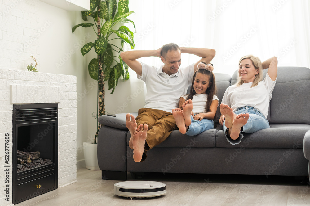 Obraz premium Young family resting on the couch while robotic vacuum cleaner doing its work at home. Household robots concept.