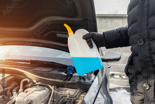 Close-up detail of driver or mechanic hand in gloves pouring blue antifreeze liquid for winter car windshield screen washing with bootle watering can. Car seasonal service and maintenance concept