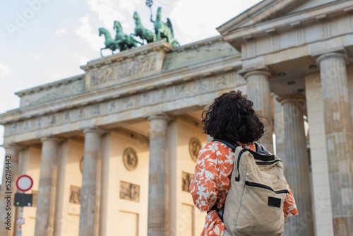 Photography back view of curly tourist with backpack standing near brandenburg gate in berlin