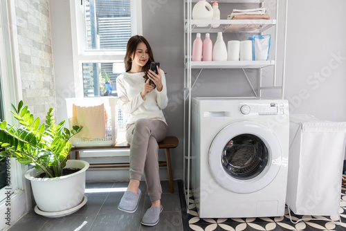 Young Asian woman sitting and using mobilephone and wait for the washing machine to finish its work