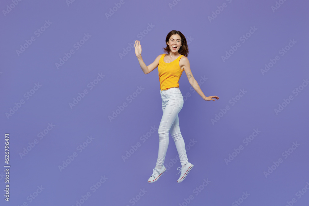 Full body side view young happy fun woman 20s she wear yellow tank shirt look camera walking go waving hand isolated on plain pastel light purple background studio portrait. People lifestyle concept.