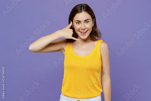 Young smiling happy fun woman 20s she wearing yellow tank shirt doing phone gesture like says call me back isolated on plain pastel light purple background studio portrait. People lifestyle concept.