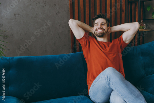 Obraz na płótnie Young minded man wears red t-shirt hold hands behind neck look aside sit on blue sofa stay at home hotel flat rest relax spend free spare time in living room indoors grey wall