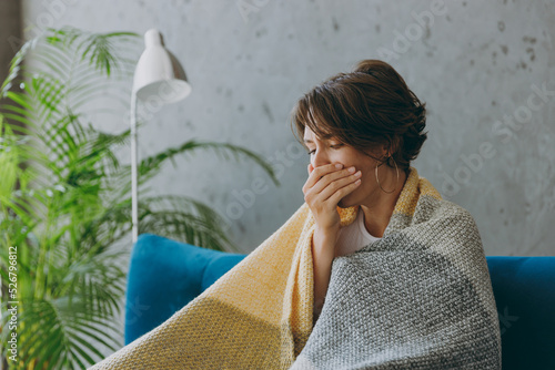 Fotografia Young sick ill sad woman wears white wrapped in plaid cough cover mouth with hand sit on blue sofa couch stay at home hotel flat spend time in living room indoors grey wall