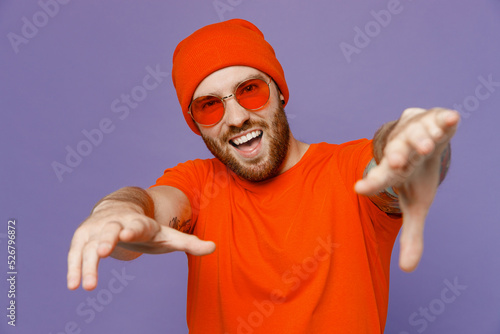 Young happy cool fun european man 20s wear red hat t-shirt sunglasses stretch hands to camera dance showoff isolated on plain pastel light purple background studio portrait People lifestyle concept. photo