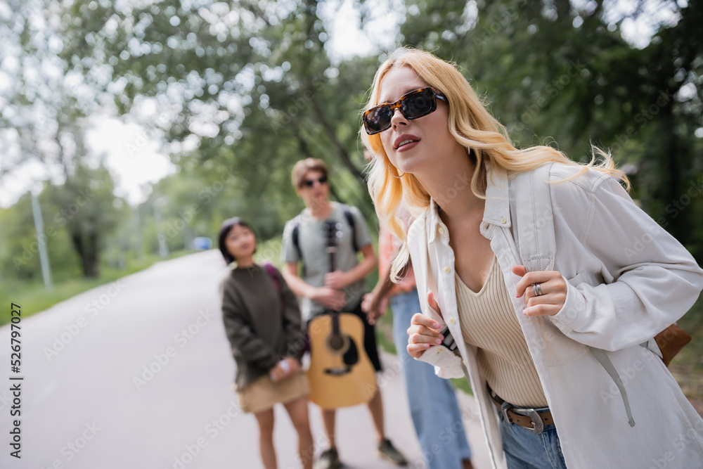 blonde woman in sunglasses near multicultural friends on blurred background.