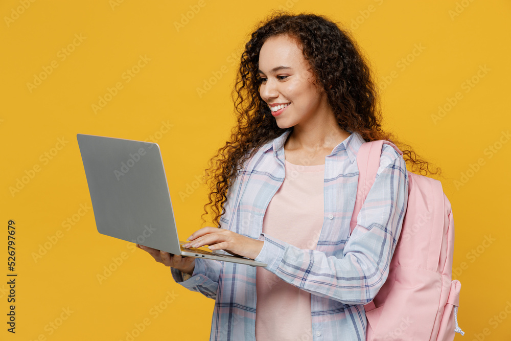 Young smiling smart black teen girl student she wear casual clothes backpack bag hold use work on laptop pc computer isolated on plain yellow color background. High school university college concept. Young smiling smart black teen girl student she wear casual clothes backpack bag hold use work on laptop pc computer isolated on plain yellow color background. High school university college concept.
