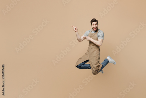 Full body young man barista barman employee in brown apron work in coffee shop jump high point finger aside on workspace isolated on plain pastel light beige background Small business startup concept.