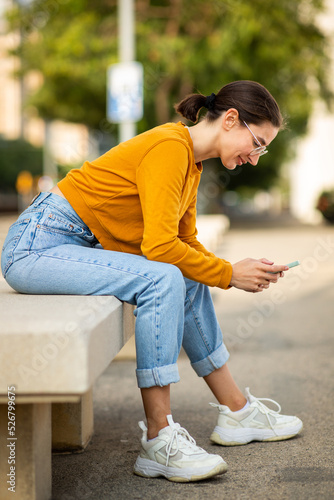 Young woman sitting on a bench outside and using her cell phone © mimagephotos