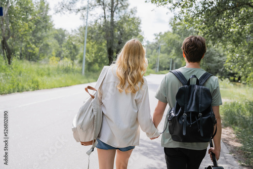 back view of young travelers holding hands and walking on road along forest.