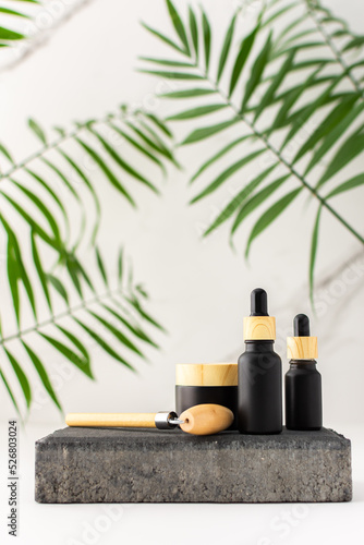 Concrete black podium for cosmetic products close-up among tropical leaves against the background of a marble white wall, bottle with a pipette and a jar of cream and a massage roller for the face