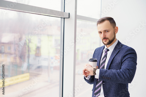 A young man in a business suit looks at his watch with a cup of coffee. A businessman in a shirt and tie and a dark jacket. A bearded man in a cafe or restaurant.