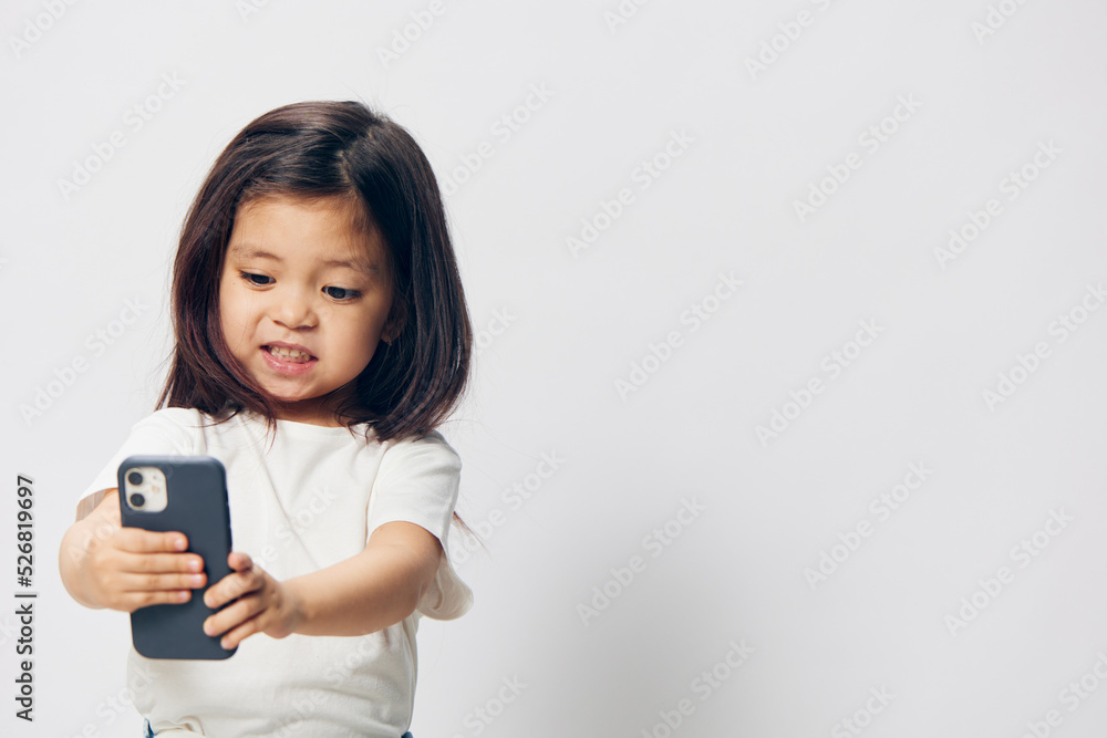 a cute little preschool girl stands on a white background in a white T-shirt and takes pictures of herself on her phone, smiling happily at the camera. The theme of children's happiness a cute little preschool girl stands on a white background in a white T-shirt and takes pictures of herself on her phone, smiling happily at the camera. The theme of children's happiness