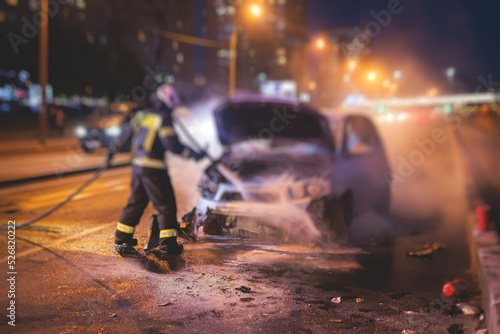 Group of fire men extinguishing and put out burning car crash after road traffic incident, fire fighting operation in the night city, firefighters with fire engine truck vehicle, emergency and rescue