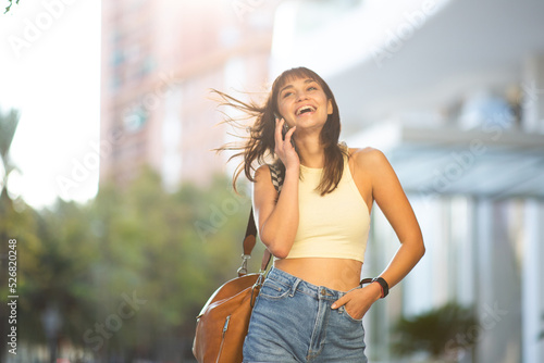 Cheerful young woman with handbag walking in the city and talking on mobile phone