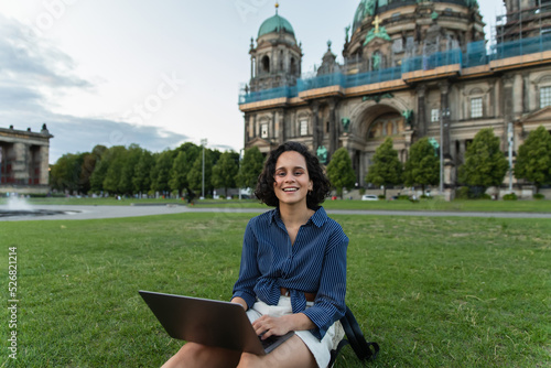 Canvas Print cheerful young woman holding laptop and sitting on grass near blurred cathedral in berlin