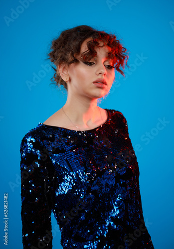 Vertical portrait of young lady standing on blue background and looking aside. Curly-haired girl in gilded dress photo