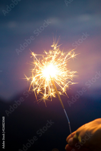 Hand holding a sparkler, or bengal fire stick, burning in outdoor setting, mountain landscape, at dusk. Holidays or magic background or wallpaper. Vertical shot.