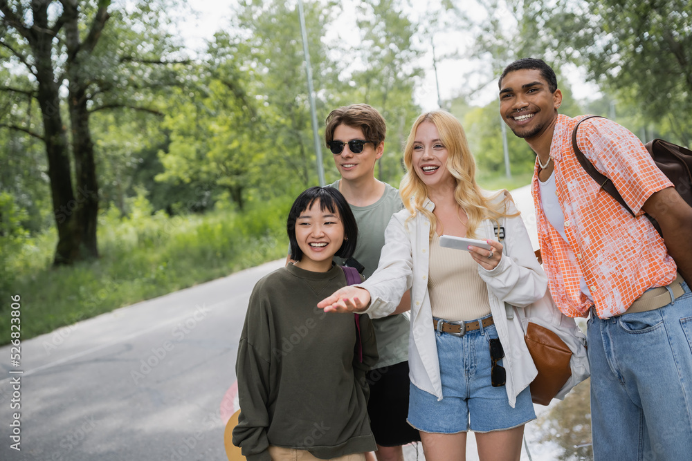 smiling woman with smartphone pointing with hand near interracial friends on road. smiling woman with smartphone pointing with hand near interracial friends on road.