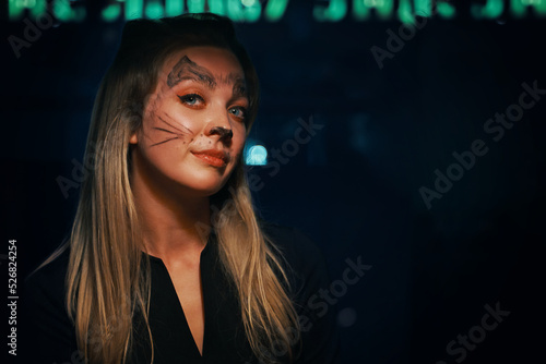 Blonde woman with cat makeup. Charming woman looks cunningly into the camera. Halloween party at a nightclub. All Saints' Night. Garland lights on the background. Photo card with copy space.