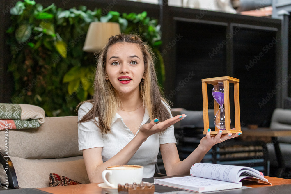 Fototapeta premium Young beautiful lady sitting at the restaurant and holding hourglass