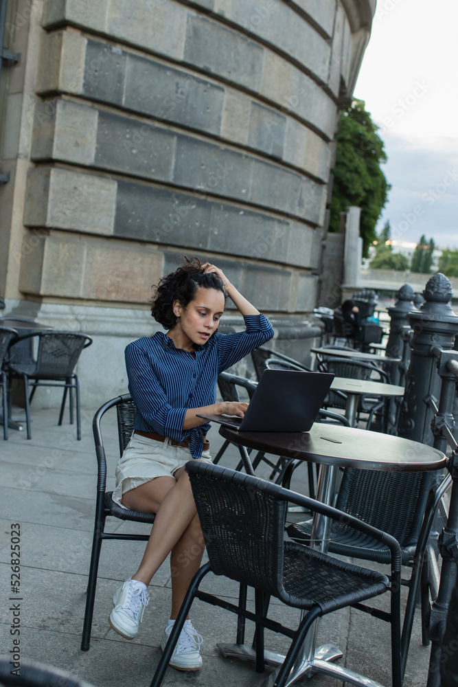 young freelancer adjusting curly hair and using laptop while sitting on summer terrace in berlin. young freelancer adjusting curly hair and using laptop while sitting on summer terrace in berlin.