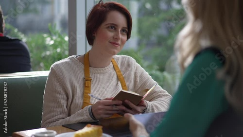 Young female staff listening to manager speak. Girl writing notes for new job sitting at table speaking with boss. Person wearing apron and holding paper and pen photo