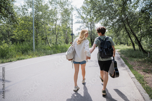 back view of young tourists with backpacks and guitar holding hands and walking on road.