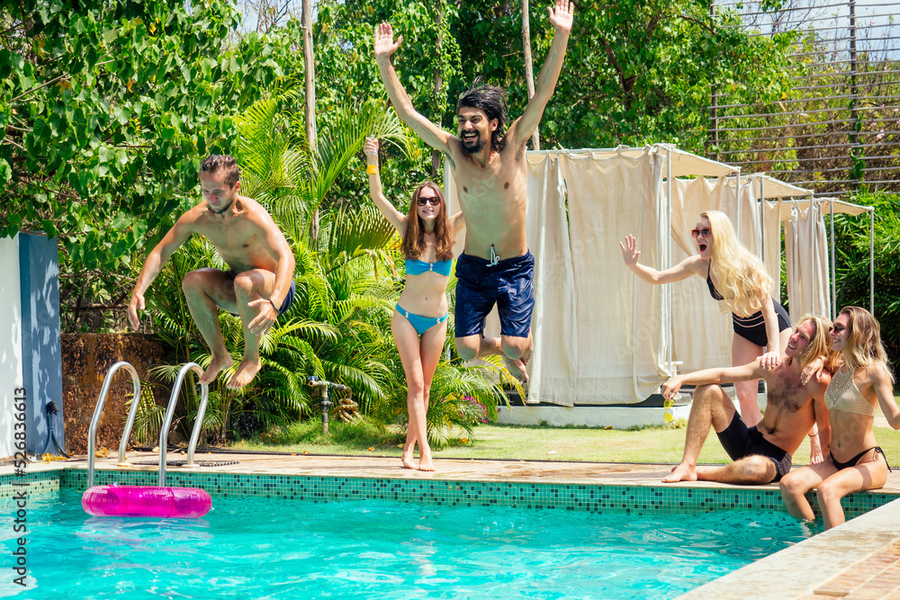 Group of happy six friends drinking jumping in pool day party with splash