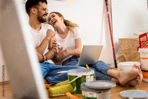 Fotografie Young couple sitting on the floor choosing color via laptop for painting the wall in their home
