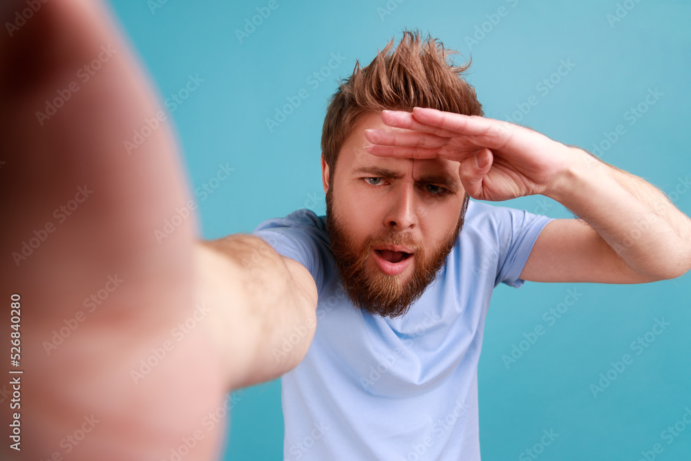 Portrait of young adult handsome curious bearded man looking into distance with attentive view, point of view, keeping hand near forehead. Indoor studio shot isolated on blue background. Portrait of young adult handsome curious bearded man looking into distance with attentive view, point of view, keeping hand near forehead. Indoor studio shot isolated on blue background.