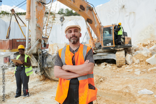 Confident foreman in reflective vest and safety helmet looking at camera while standing on construction site against caterpillar truck