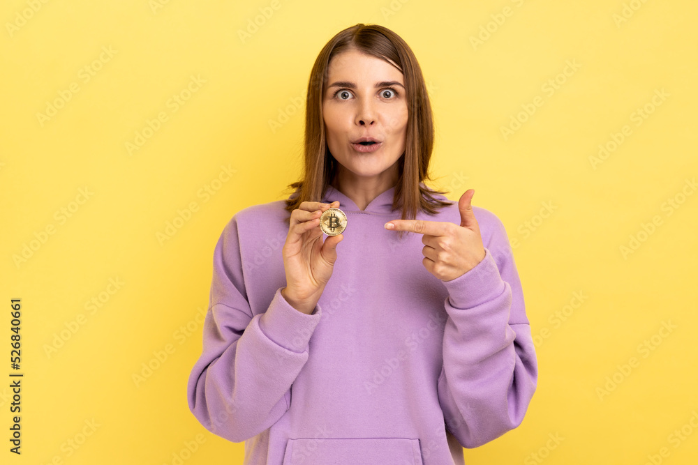 Cryptocurrency, digital money. Surprised excited woman showing golden bitcoin and looking with amazement, pointing at btc coin, wearing purple hoodie. Indoor studio shot isolated on yellow background. Cryptocurrency, digital money. Surprised excited woman showing golden bitcoin and looking with amazement, pointing at btc coin, wearing purple hoodie. Indoor studio shot isolated on yellow background.