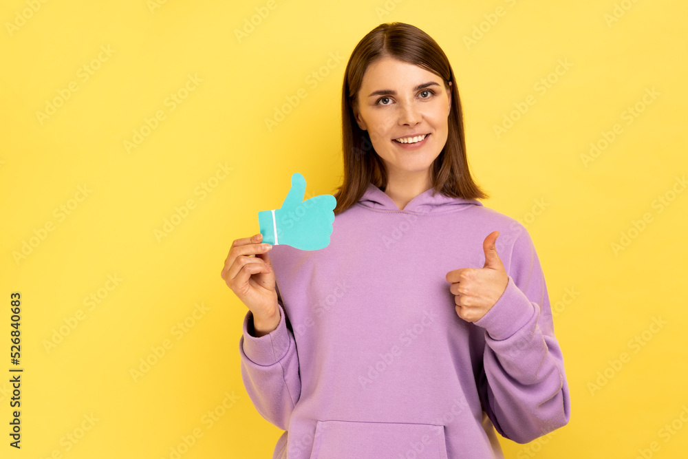 Smiling woman showing thumbs up blue paper sign, liking and recommending posts in social networks, asking to rate, wearing purple hoodie. Indoor studio shot isolated on yellow background. Smiling woman showing thumbs up blue paper sign, liking and recommending posts in social networks, asking to rate, wearing purple hoodie. Indoor studio shot isolated on yellow background.