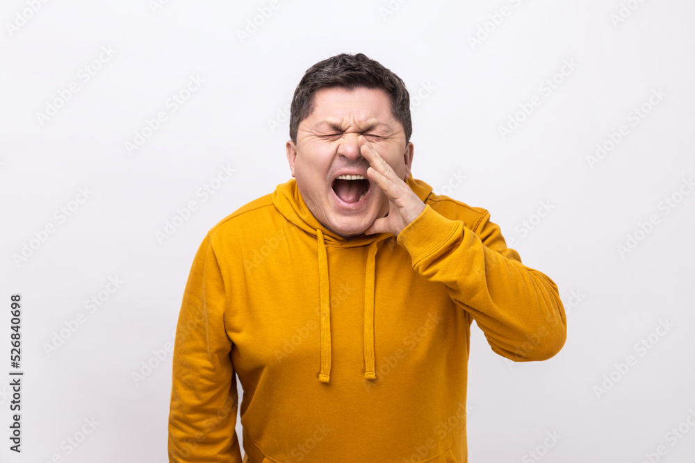 Angry nervous middle aged man loudly yelling widely opening mouth holding hands on face, screaming announcing his opinion, wearing urban style hoodie. Indoor studio shot isolated on white background. Angry nervous middle aged man loudly yelling widely opening mouth holding hands on face, screaming announcing his opinion, wearing urban style hoodie. Indoor studio shot isolated on white background.