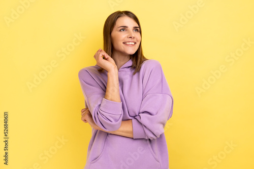 Portrait of adorable dreamy young woman standing and dreaming, making wish, love, looking away with toothy smile, wearing purple hoodie. Indoor studio shot isolated on yellow background. © khosrork