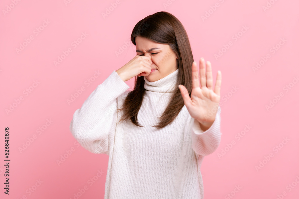Woman grabbing nose, grimacing in disgust and showing stop gesture, expressing repulsion to stink, wearing white casual style sweater. Indoor studio shot isolated on pink background. Woman grabbing nose, grimacing in disgust and showing stop gesture, expressing repulsion to stink, wearing white casual style sweater. Indoor studio shot isolated on pink background.