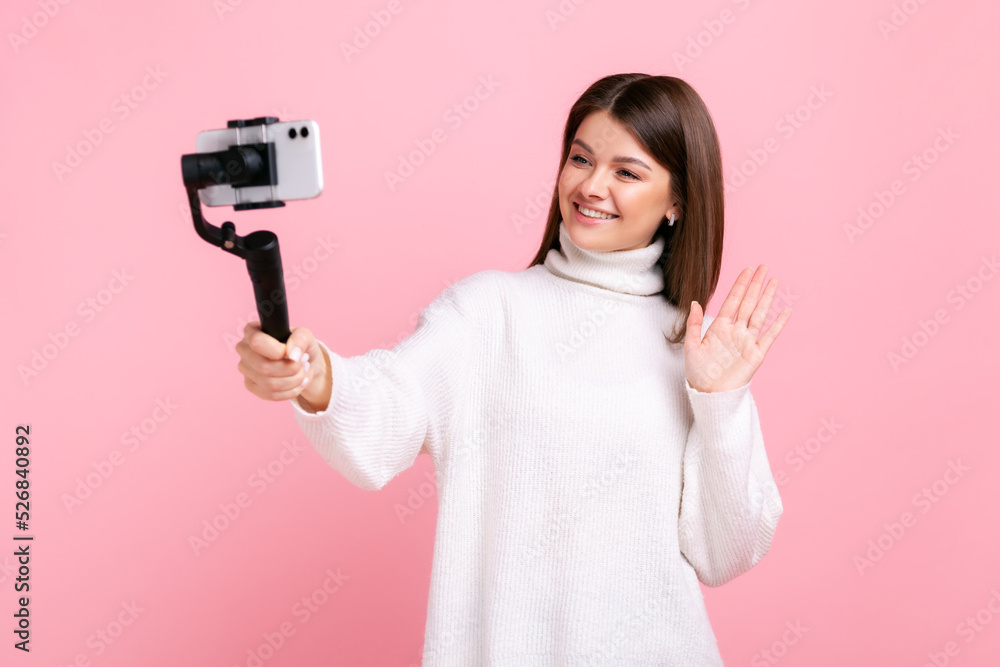 happy woman blogger using smart phone and steadicam for streaming, waving hand, greeting followers, wearing white casual style sweater. Indoor studio shot isolated on pink background. happy woman blogger using smart phone and steadicam for streaming, waving hand, greeting followers, wearing white casual style sweater. Indoor studio shot isolated on pink background.