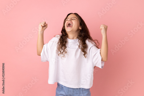 Portrait of little girl wearing white T-shirt expressing winning gesture with raised fists and screaming, celebrating victory. Indoor studio shot isolated on pink background.