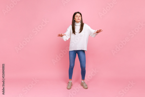 Full length portrait of disoriented girl walks with eyes closed, holding out her hands to find road, wearing white casual style sweater. Indoor studio shot isolated on pink background.