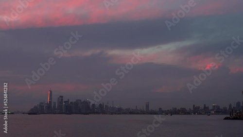 Timelapse of getting dark above the Manhattan skyscrapers in the evening seen from the American Veterans Memorial Pier. Sailing ferries. Clouds are lightened with pink and purple by the sun. USA, NYC