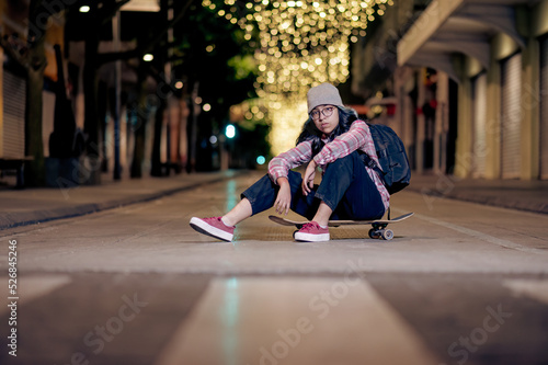 Attractive young female skater in a hat and backpack sitting on a skateboard on the night city street looking at a camera