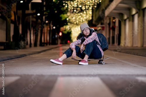 Attractive young female skater in hat and backpack sitting on a skateboard on the night city street.