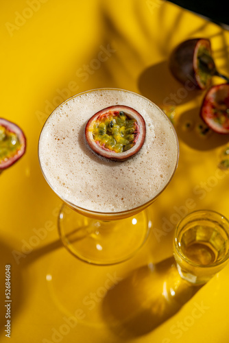 Glass of Porn Star Martini cocktail with passionfruit on yellow table with dark background, raw fruits around. Summer exotic fruit alcohol refreshing drink with foam. Copy space.
