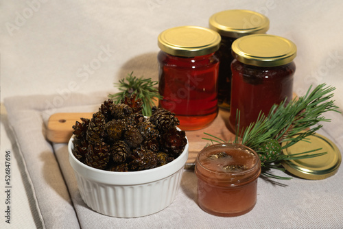 On a light fabric background on a napkin is delicious caramel jam and syrup from young small pine cones in a glass jar and a bowl with branches and needles