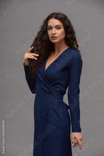 Young glamorous female fashion model in navy blue smart casual dress touching her dark long wavy hair while looking at camera