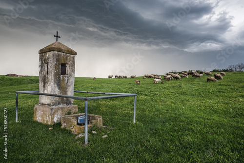 Small roadside shrine on a meadow in Banat region in Vojvodina, Serbia. Behind, under the stormy skies, a flock of sheep and goats are grazing grass up on the hill. photo